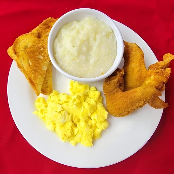 A white plate with scrambled eggs, grits, toast, and fried chicken wings on a red background.